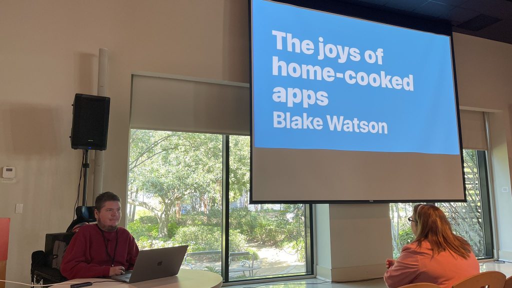 Photo of me at MagnoliaJS. I’m sitting in my wheelchair at a small table by the podium. My laptop is open and off to the side behind me is my presentation slideshow. The cover slide reads "The joys of home-cooked apps. Blake Watson."