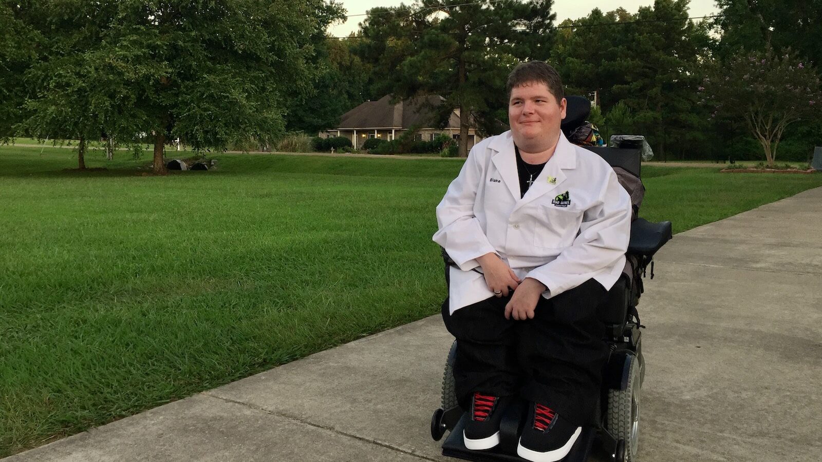 Photo of me sitting in my wheelchair in the driveway outside my house wearing a lab coat.